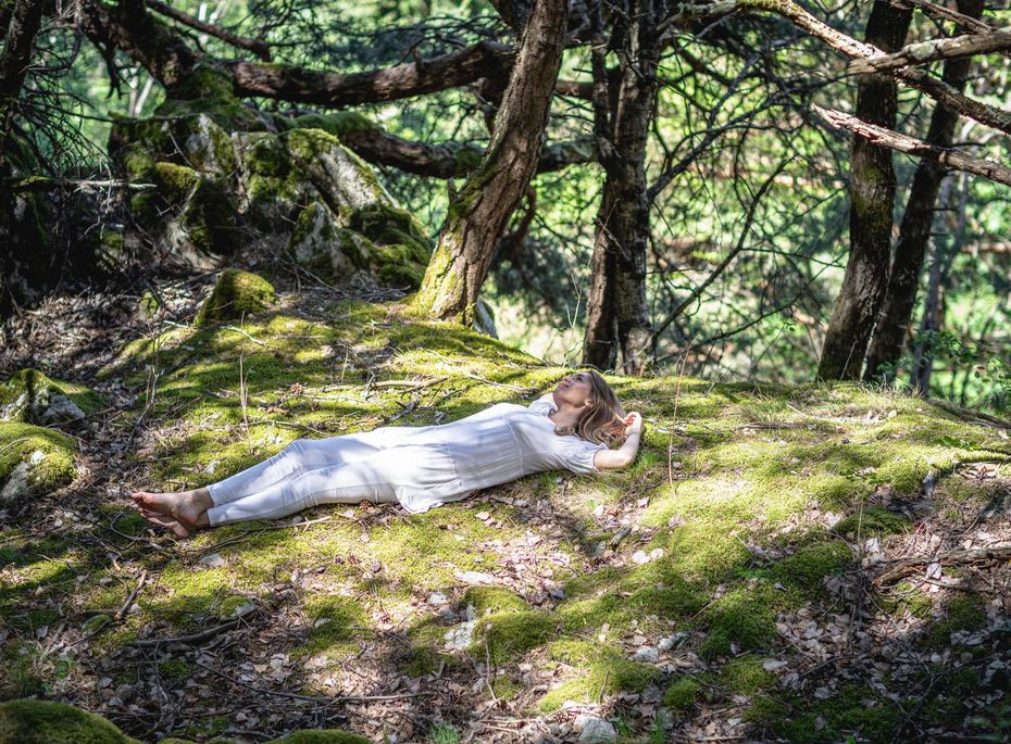 Eine Frau liegt in heller Kleidung entspannt auf einer moosigen Waldlichtung zwischen Bäumen.