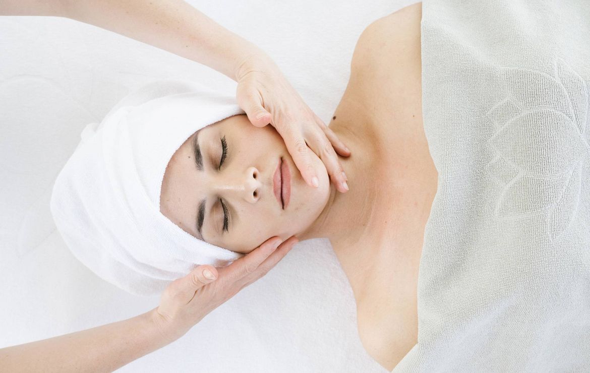 A therapist gently massages a woman's face during a facial treatment on a white treatment bed.