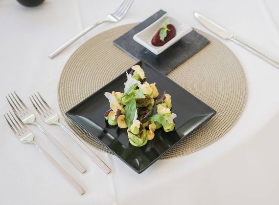 A black square plate holds a composed healthy gourmet dish with a small sauce cup on a round placemat.