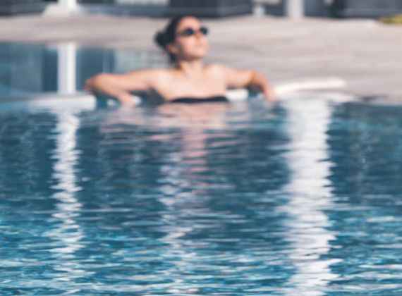 A woman in sunglasses relaxes at the edge of an outdoor pool with calm blue water.