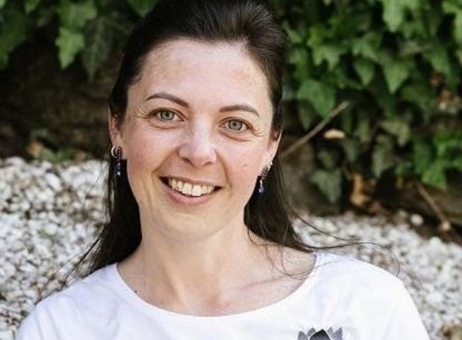 Eine lächelnde Frau im weißen T-Shirt sitzt vor einer grünen Blattwand und schaut in die Kamera.