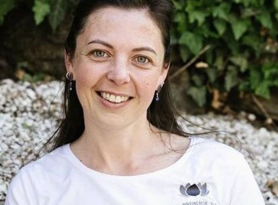 Porträt einer Mitarbeiterin im Preidlhof Eine lächelnde Frau im weißen T-Shirt sitzt vor einer grünen Blattwand und schaut in die Kamera.