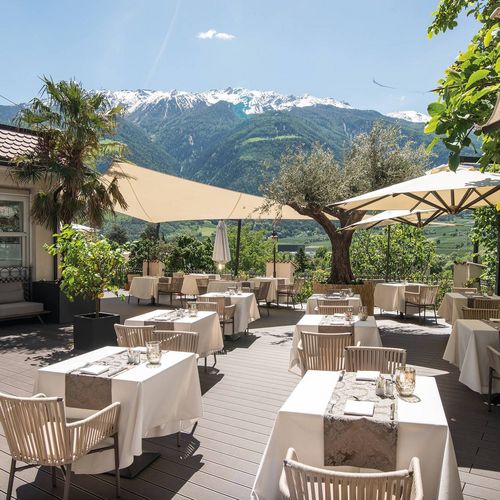 Eine gedeckte Restaurantterrasse mit Tischen, Sonnenschirmen und Blick auf die Berge.