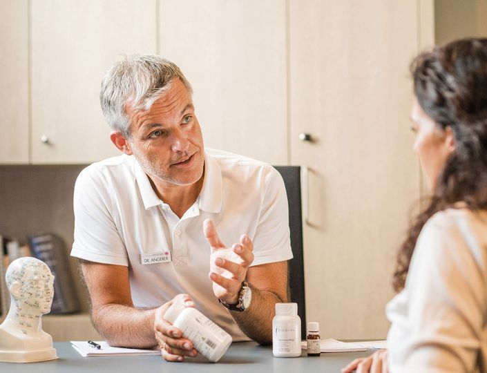Ein Mitarbeiter berät eine Frau am Tisch und zeigt ihr ein Pflegeprodukt in einem Behandlungsraum.