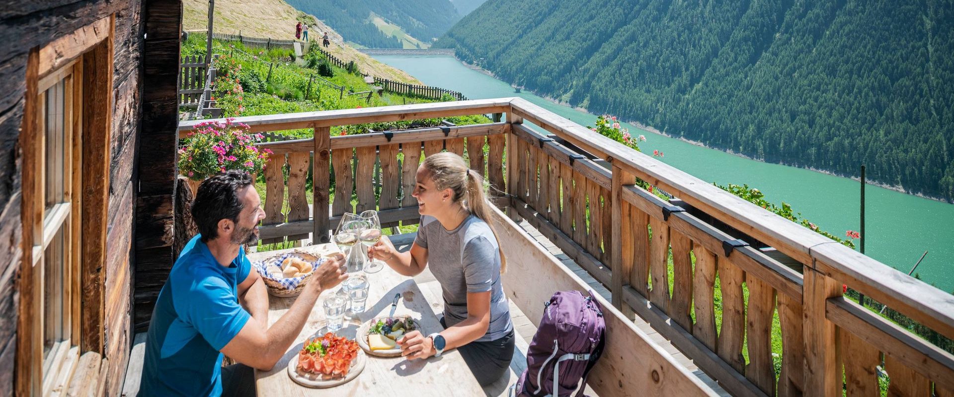 Ein Paar isst auf einer Holzterrasse mit Blick auf ein Tal, einen See und die umliegenden Berge.