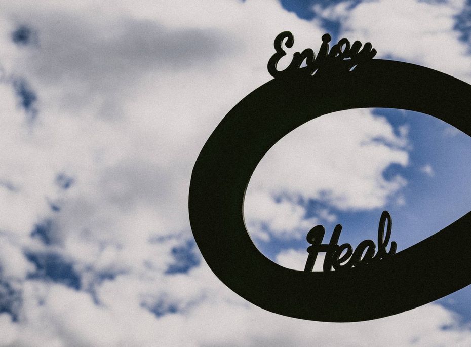 Ein schwarzer, ovaler Schriftzug mit den Worten „Enjoy“ und „Heal“ vor wolkigem Himmel.