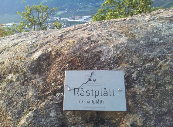 Eine Metalltafel mit der Aufschrift "Rastplatt" ist auf einem großen Felsen befestigt, im Hintergrund liegt das Tal.
