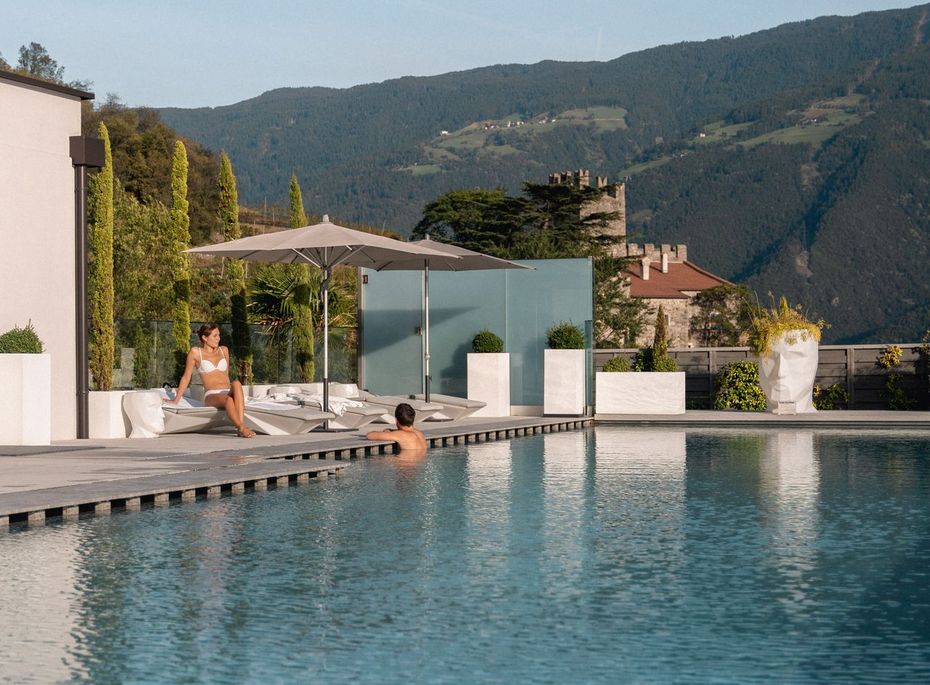 Infinity Pool im Preidlhof in Naturns Ein Infinity-Pool mit Liegen und Sonnenschirm vor Bergkulisse, am Beckenrand sitzen zwei Gäste.