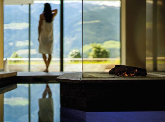 A woman stands by the window in a sauna room, with mountains visible outside and a pool reflection in the foreground.