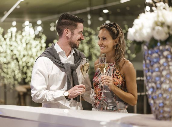 Ein lächelndes Paar hält im eleganten Innenbereich zwei Gläser Champagner und blickt sich an.