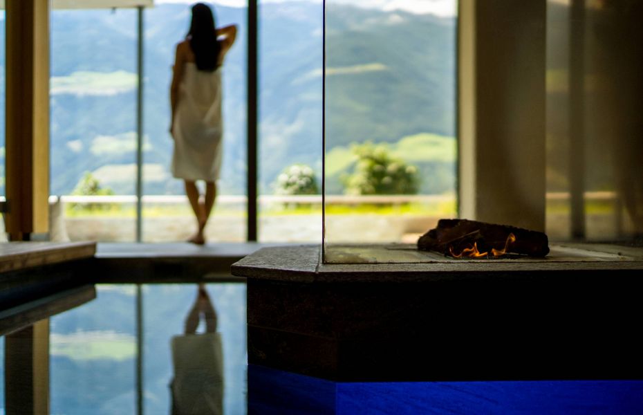 Sauna im Wellnesshotel in Südtirol Eine Frau steht mit Blick auf die Berge in einem hellen Spa-Bereich mit Wasserbecken und Feuerstelle.