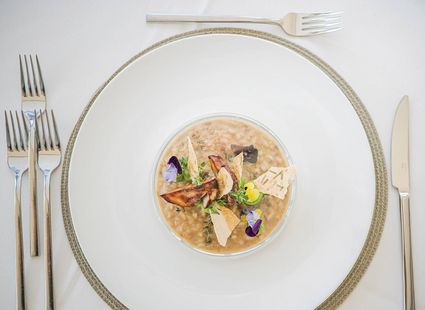 Ein feines Risotto mit Gemüse und essbaren Blüten liegt auf einem großen weißen Teller mit Silberbesteck.