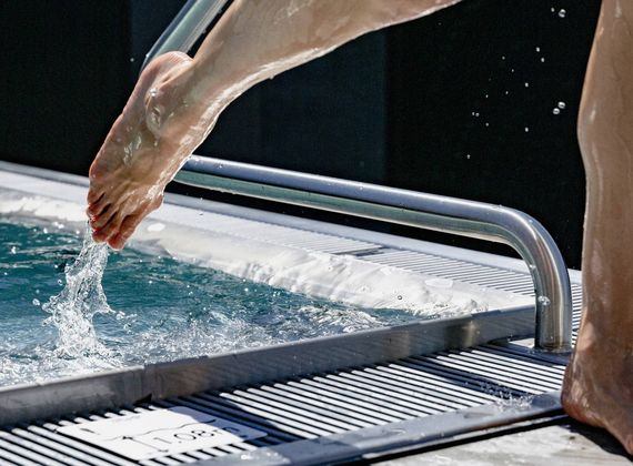 A person lowers one leg into a thermal spa pool beside a metal ladder and wet deck.
