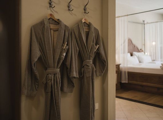 Zwei Bademäntel hängen an Wandhaken; im Hintergrund ist ein helles Hotelzimmer mit Bett zu sehen.
