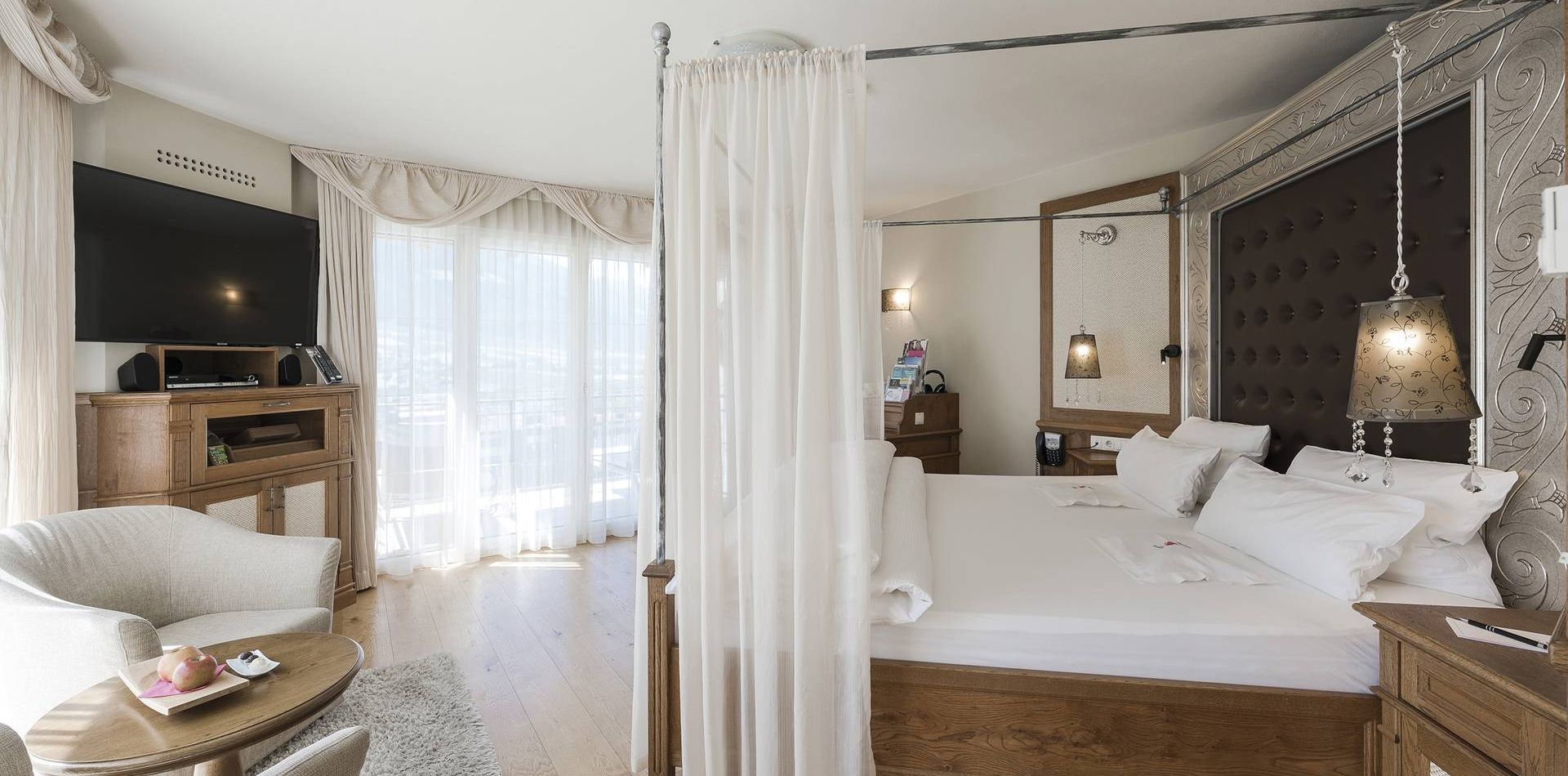 A luxury suite bedroom with a four-poster bed, sheer curtains, armchairs and a TV in a bright hotel room.