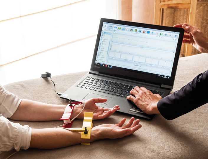 Eine Person misst mit Elektroden am Arm, während am Laptop eine medizinische Auswertung angezeigt wird.