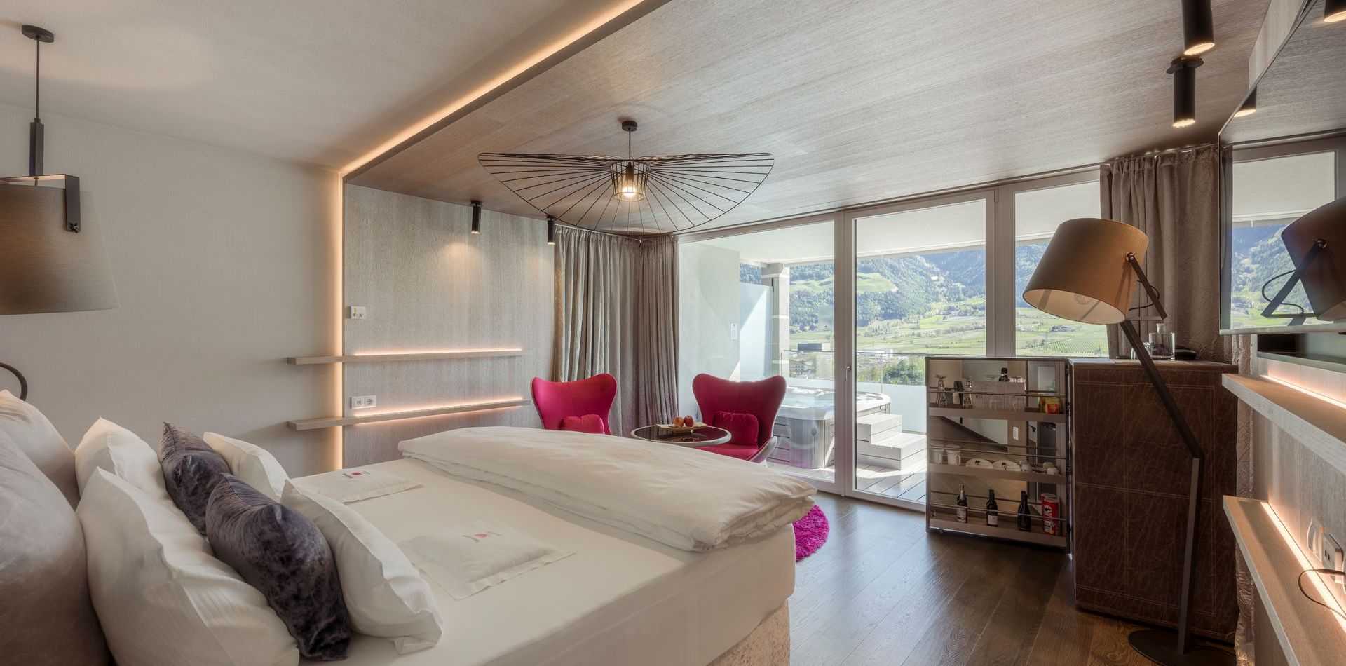 Eine helle Luxussuite mit großem Bett, rotem Sessel, Holzdetails und Blick auf die Berge.