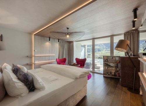 Eine helle Luxussuite mit großem Bett, rotem Sessel, Holzdetails und Blick auf die Berge.