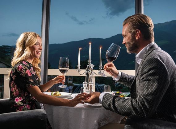 Ein Paar sitzt bei einem romantischen Dinner am Tisch und hält Weingläser vor der Abendlandschaft.