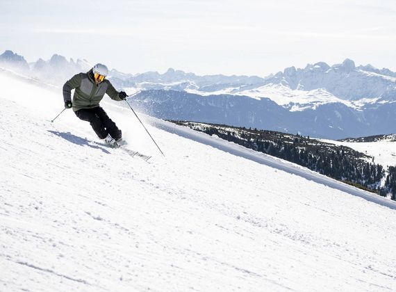 A skier carves down a snowy alpine slope with mountain peaks in the background.