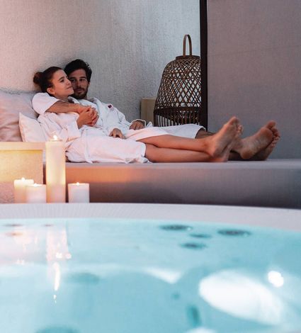 A couple in white robes relaxes on a lounge bed beside a softly lit spa pool.