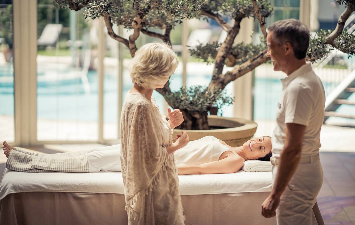 A woman lies on a treatment bed while two people stand nearby in a spa room with poolside views.