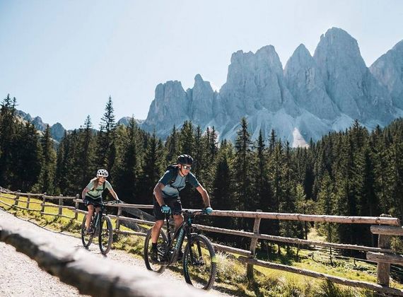 Zwei Mountainbiker fahren auf einem Weg mit Holzzaun vor felsigen Gipfeln und Nadelwald.