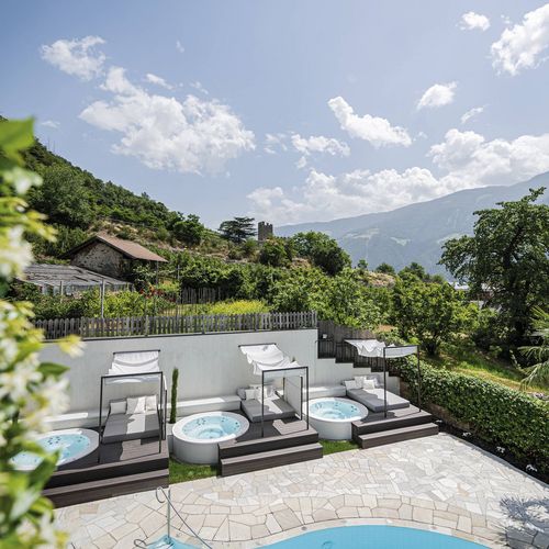 Ein Außenpool mit Liegen und kleinen Whirlpools vor grüner Hügellandschaft und Bergpanorama.