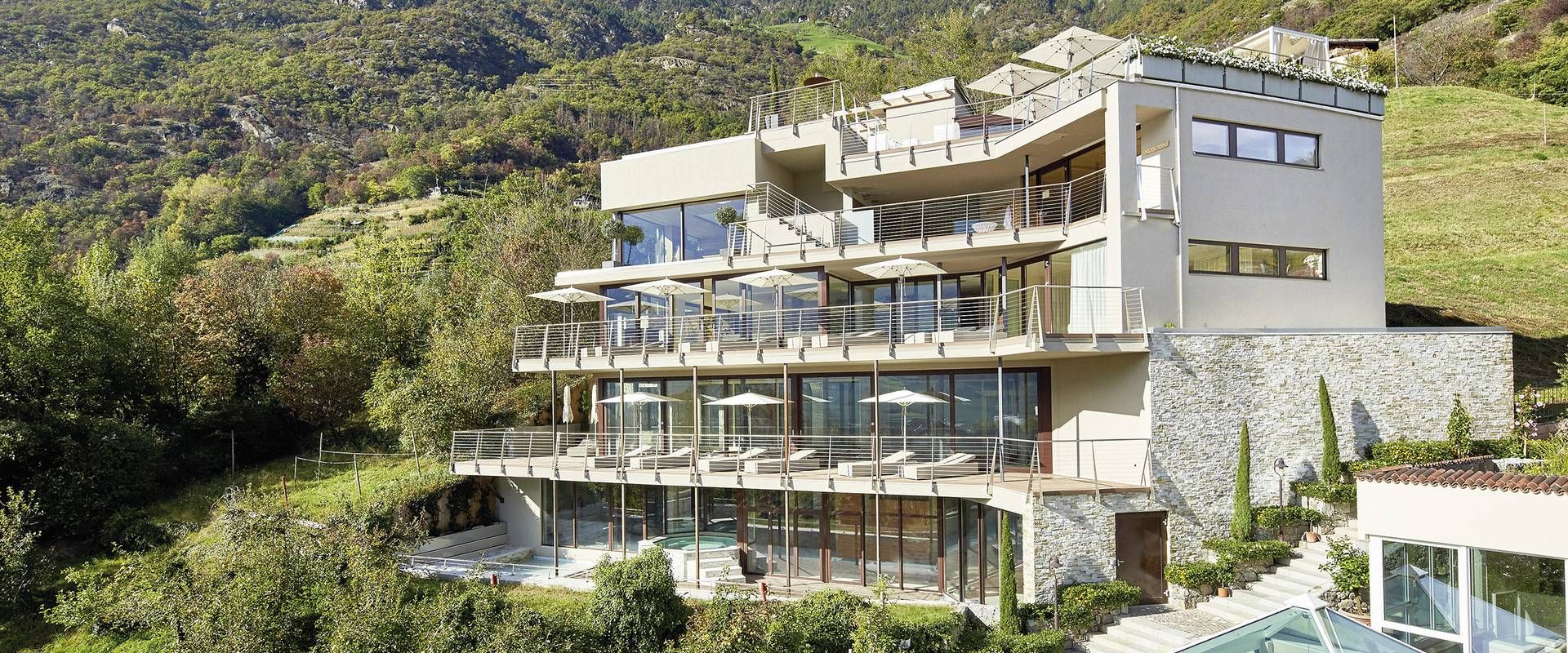 Das Wellnesshotel am Hang zeigt mehrere Terrassen mit Glasfronten vor grünen Hügeln.