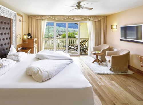 A bright luxury suite with a large bed, seating area, TV, and sliding doors to a balcony with mountain views.