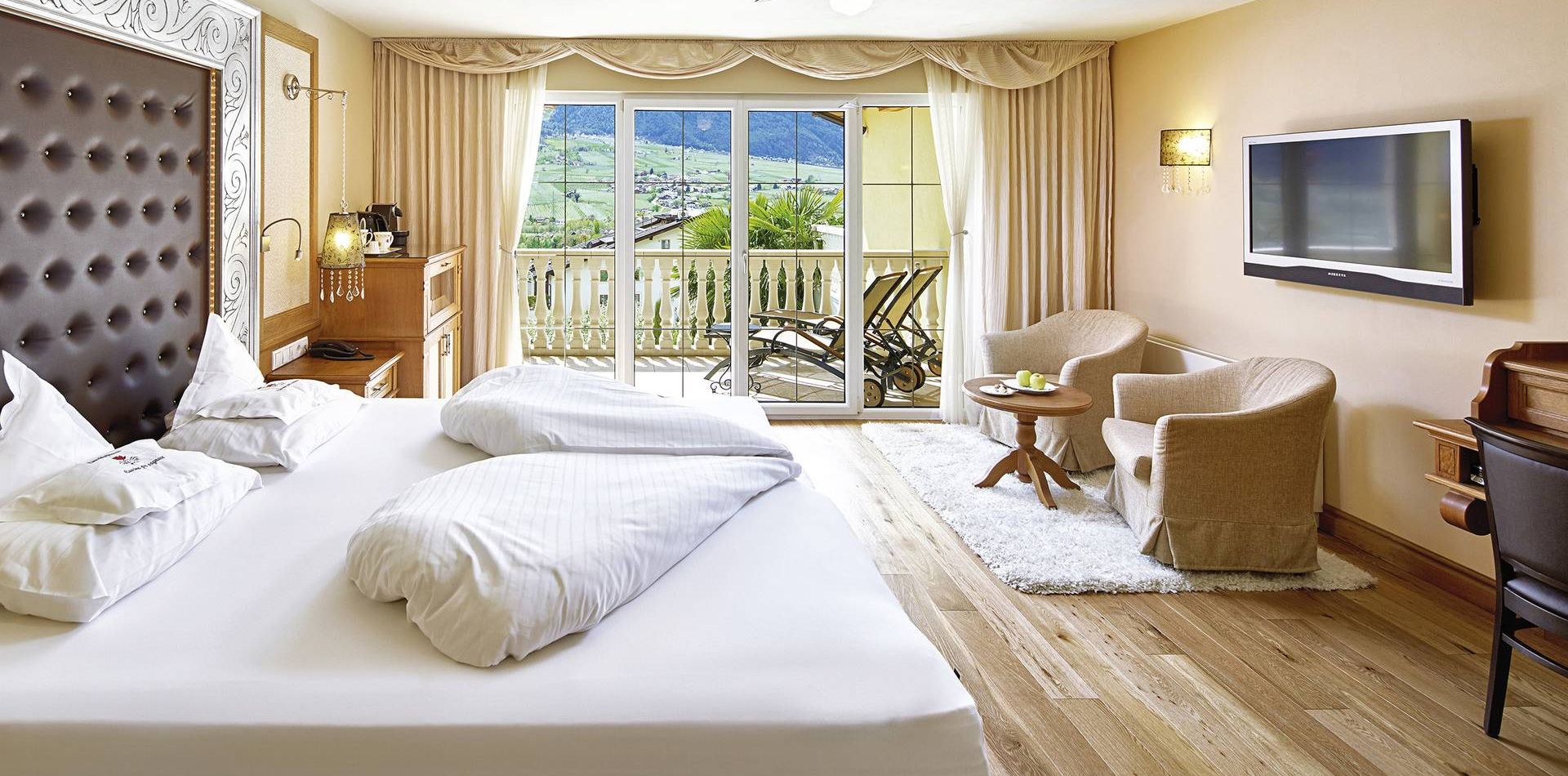 A bright luxury suite with a large bed, seating area, TV, and sliding doors to a balcony with mountain views.