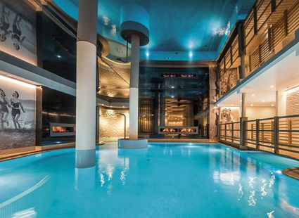 Ein großzügiger Indoor-Pool mit türkisblauem Wasser in einer modern gestalteten Wellnesshalle.