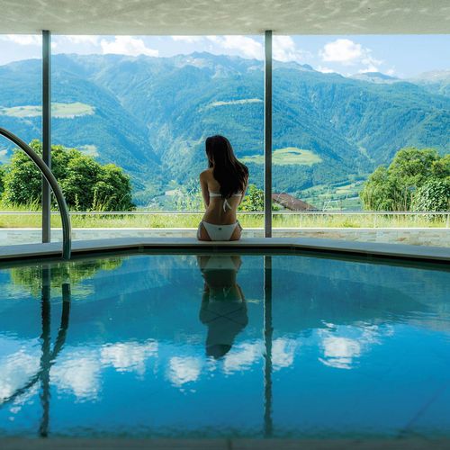 Eine Frau sitzt am Infinity Pool vor einer großen Glasfront mit Blick auf die Berglandschaft.