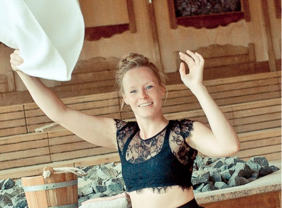 Eine Frau schwenkt ein Tuch in der Sauna neben einem Holzeimer und einem Becken mit Steinen.