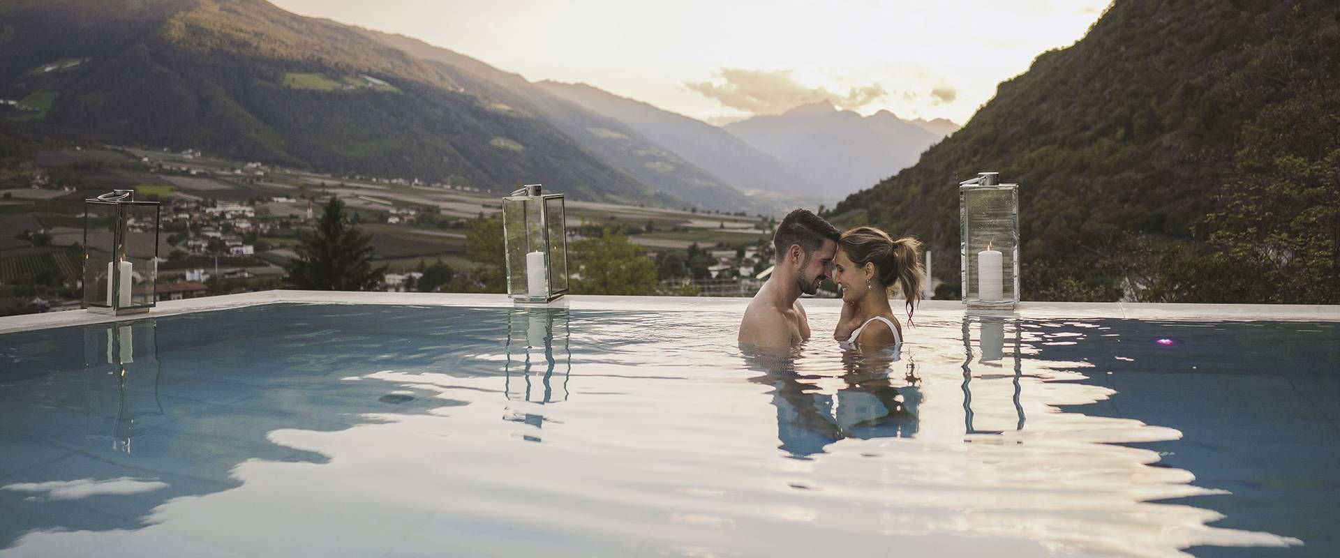 Ein Paar steht in einem Infinity-Pool vor einer Bergkulisse im Abendlicht.