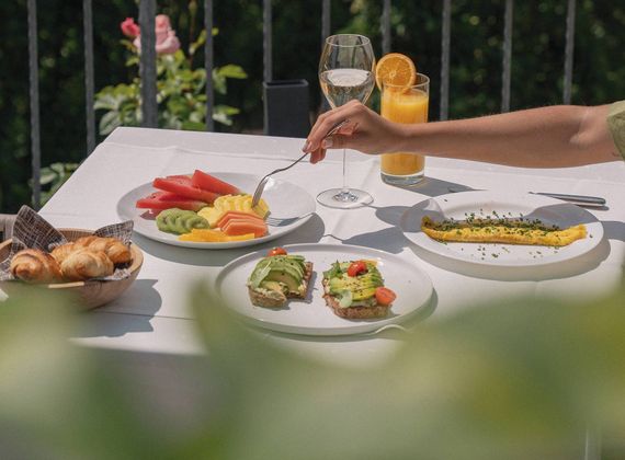 A breakfast table on a terrace with fruit, croissants, avocado toast, an omelette, juice and a glass of wine.