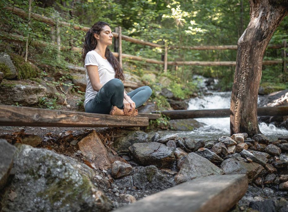 Wellness-Ruheplatz im Grünen am Bach Eine Frau sitzt auf einer Holzbank am Bach und meditiert im Grünen zwischen Steinen und Bäumen.