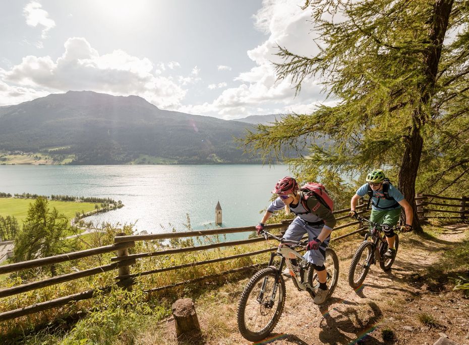 Mountainbiken am See bei Naturns Zwei Mountainbiker fahren auf einem Waldweg oberhalb eines Sees mit Bergen im Hintergrund.
