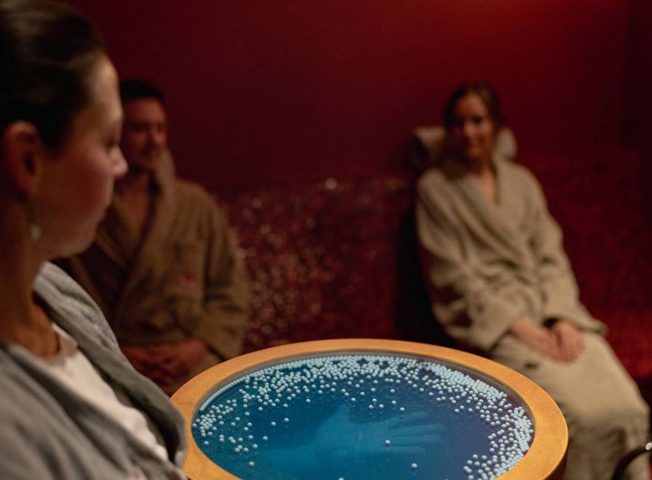 Rasul-Zeremonie im Wellnesshotel in Naturns Eine Mitarbeiterin hält eine runde Schale mit blauem Inhalt, während drei Gäste in Bademänteln im Ruheraum sitzen.