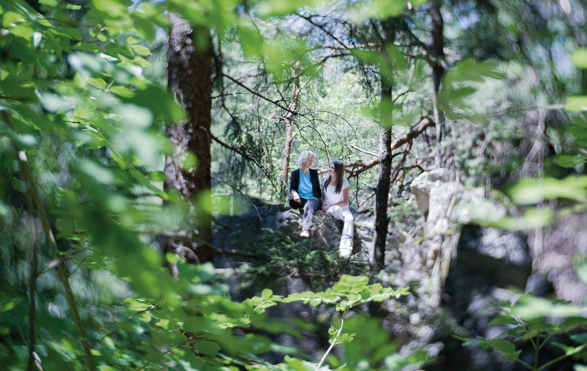 Zwei Frauen sitzen auf einem Felsen im Grünen und genießen eine Wanderpause im Wald.