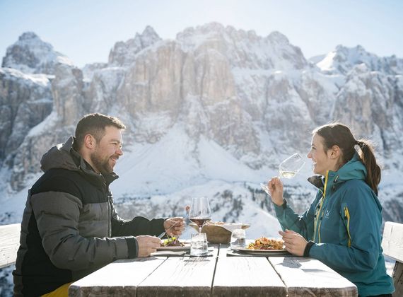 Ein Paar sitzt im Freien an einem Holztisch und isst vor einer verschneiten Bergkulisse im Winterurlaub in Südtirol.