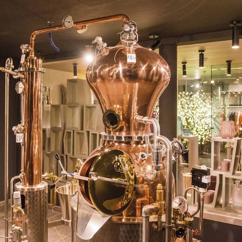 Eine kupferne Destillieranlage steht in einem stilvollen Innenraum mit Regalen und Glasflächen im Hintergrund.