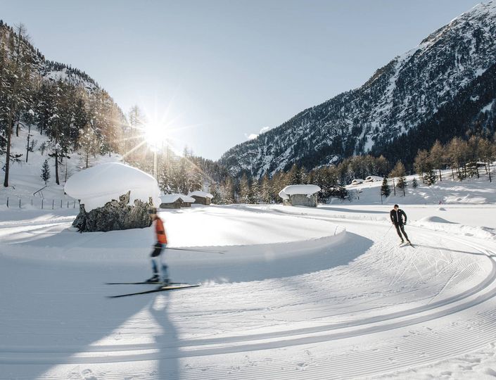 Zwei Skifahrer gleiten auf einer präparierten Loipe durch eine verschneite Berglandschaft im Sonnenlicht.