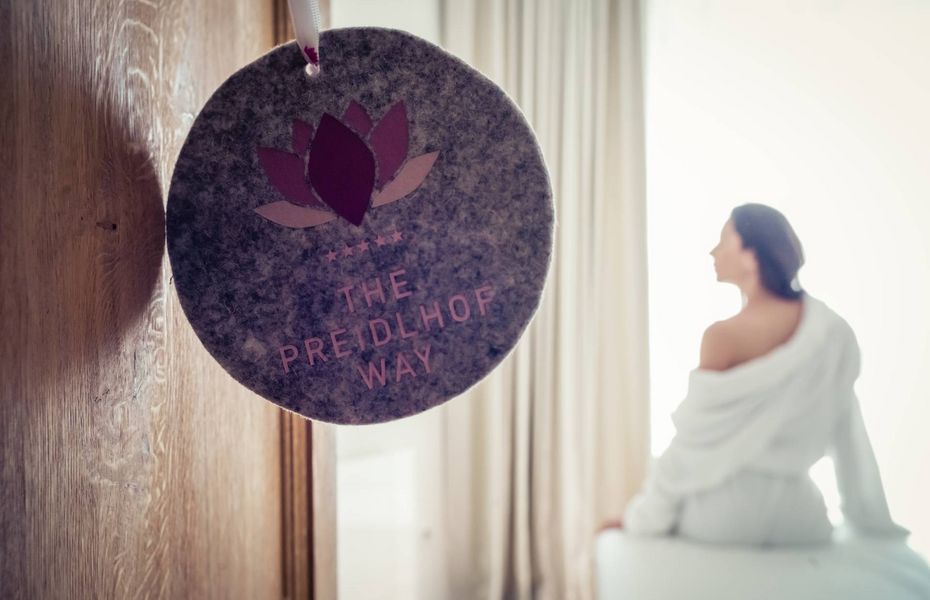 Wellnessbereich im Preidlhof mit Ruheliege Eine Frau sitzt im Bademantel auf einer Ruheliege neben einem hängenden Preidlhof-Schild im hellen Wellnessbereich.
