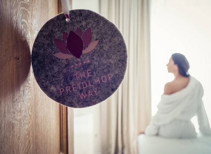 Eine Frau sitzt im Bademantel auf einer Ruheliege neben einem hängenden Preidlhof-Schild im hellen Wellnessbereich.
