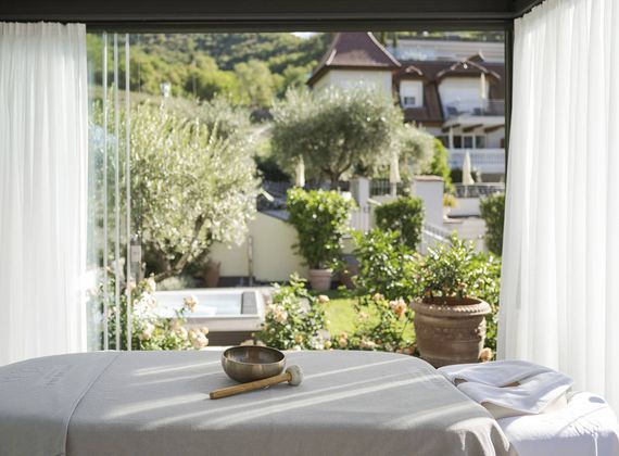 Un lettino per trattamenti è rivolto verso il giardino luminoso attraverso ampie vetrate, con una ciotola spa e teli sul tavolo.
