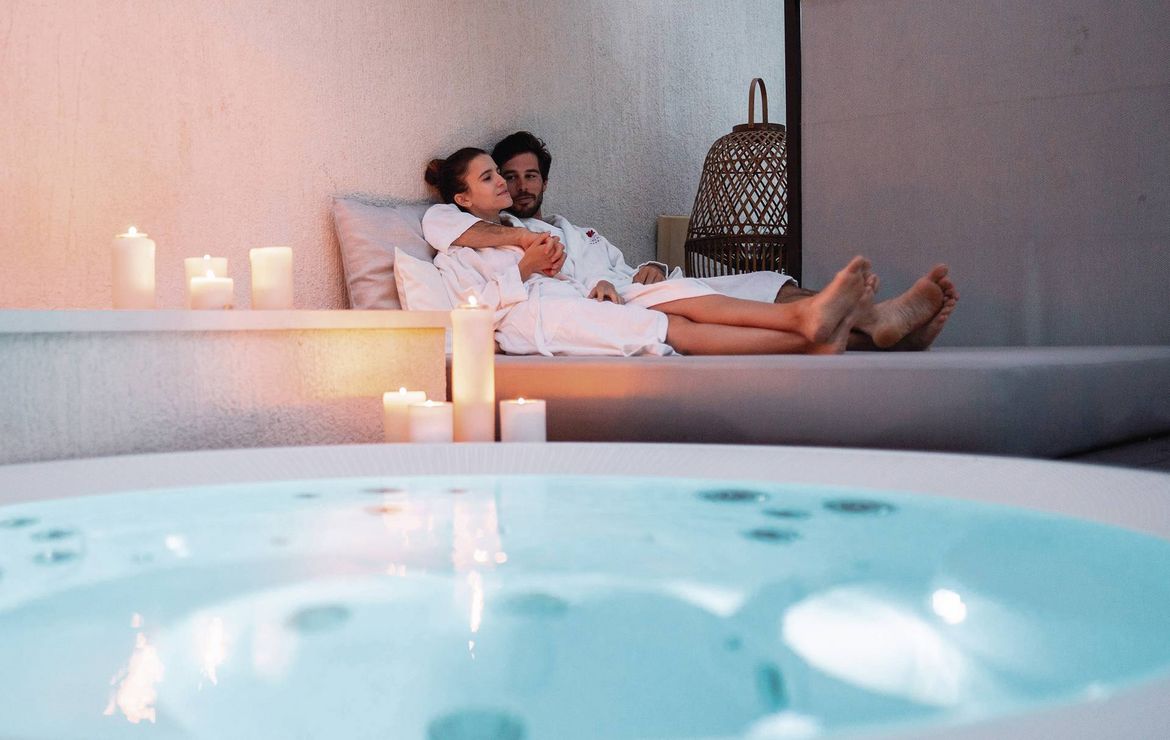 A couple relaxes on a daybed beside an indoor spa pool with candles and soft lighting.