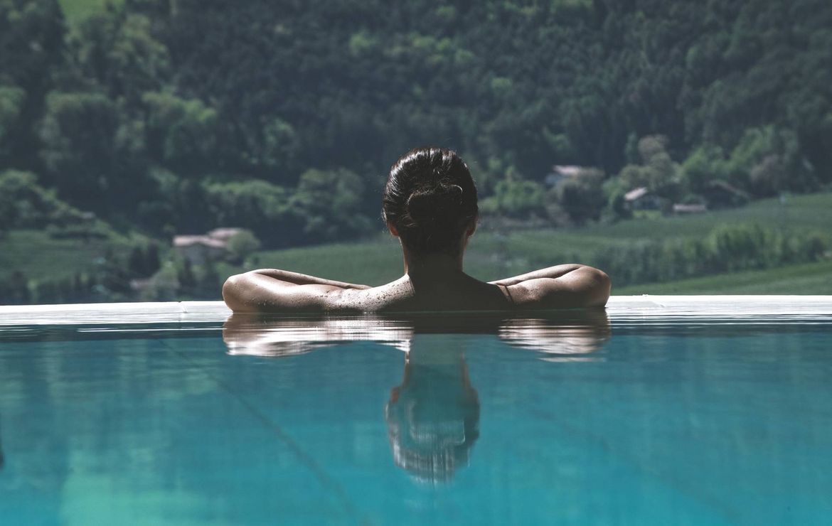 Una persona riposa con le braccia sul bordo di una piscina a sfioro affacciata su una valle verde.