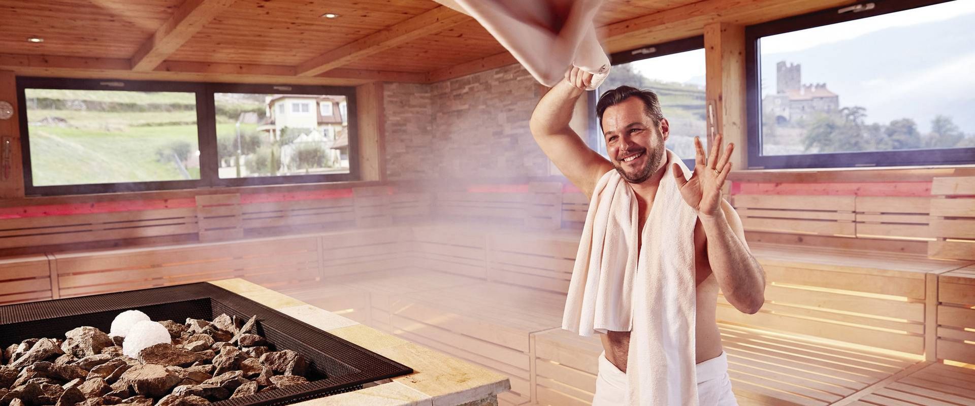 Ein Mann steht mit einem Handtuch in einer Holzsauna und begrüßt den Aufgussbereich.
