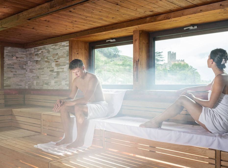 Sauna im Preidlhof in Naturns Ein Mann und eine Frau sitzen in einer Holzsauna auf weißen Liegen und blicken durch die Fenster nach draußen.